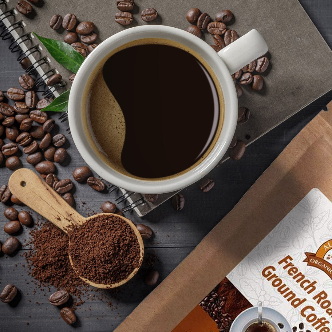 A cup of Alex's Low-Acid Organic Coffee™ - French Roast Fresh Ground (12oz) by Alex's Low Acid Coffee sits on a notebook, surrounded by coffee beans, a wooden scoop with ground coffee, and the product bag on a dark wooden surface.
