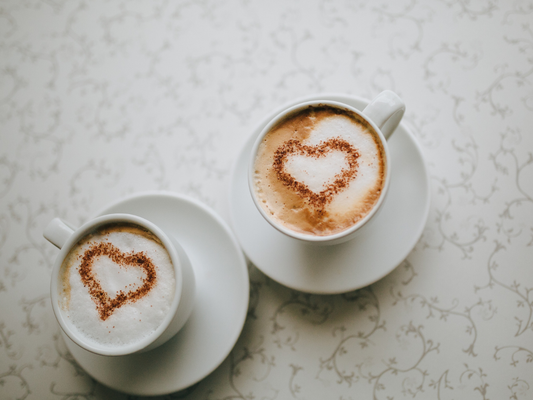 Two cups of coffee on a light surface with heart-shaped foam art on top.