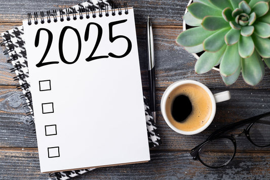A notepad labeled "2025" with checkboxes sits on a wooden table beside a cup of coffee, a pen, a pair of glasses, and a small succulent plant.