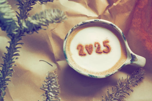 A close-up of a cup of cappuccino topped with cocoa powder forming the numbers "2025," surrounded by brown paper and evergreen branches.
