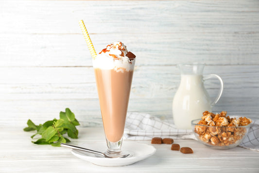A tall glass filled with chocolate milkshake topped with whipped cream and a drizzle of chocolate sits on a plate with a spoon, accompanied by a pitcher of milk and a bowl of caramel popcorn on a light wooden surface.