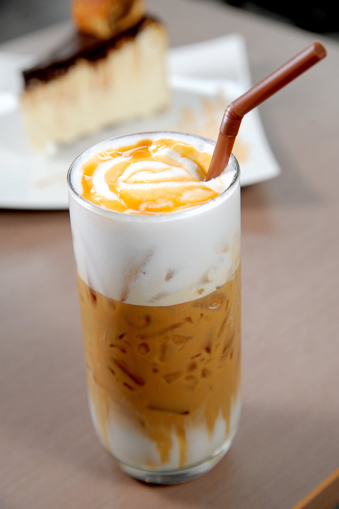 A tall glass of iced coffee with a swirl of cream and caramel on top, accompanied by a brown straw, sits on a table with a slice of cake in the background.