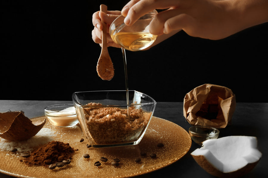 A hand pours liquid into a glass bowl filled with brown sugar, surrounded by various ingredients like coconut, coffee beans, and a small bowl of white powder on a textured surface.
