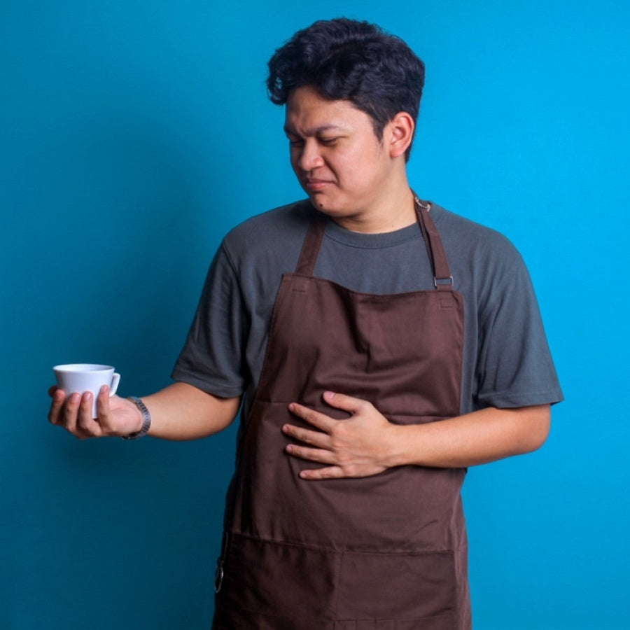 A man in a brown apron stands against a blue background, holding a white cup in one hand while grimacing and clutching his stomach with the other, suggesting discomfort.