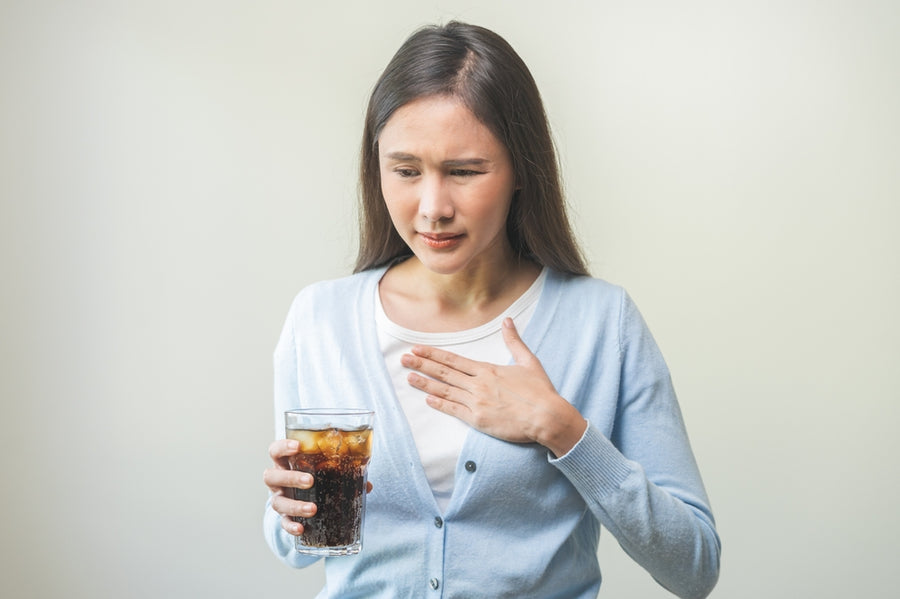 A woman in a light blue cardigan holds a glass of cola and looks concerned, with one hand on her chest, suggesting discomfort or distress in a neutral setting.