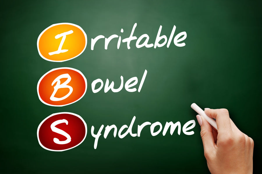 A hand holding a piece of chalk writes "Irritable Bowel Syndrome" on a green chalkboard, with the letters "IBS" highlighted in orange, red, and white.