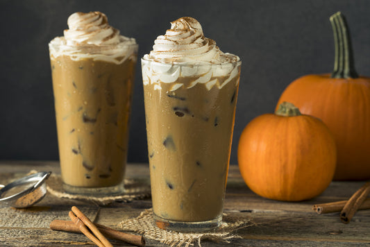 Two tall glasses of iced coffee topped with whipped cream and cinnamon sit on a rustic wooden table, accompanied by whole pumpkins and cinnamon sticks, creating a cozy autumn atmosphere.