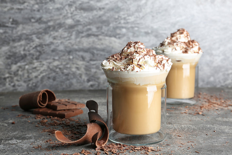 Two clear glasses filled with creamy coffee topped with whipped cream and chocolate shavings sit on a textured gray surface, surrounded by chocolate curls and shavings.