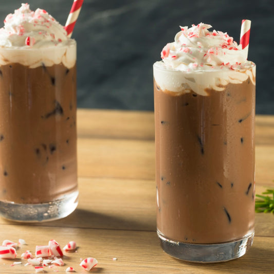 Two tall glasses of iced chocolate drinks topped with whipped cream and crushed peppermint sit on a wooden table, with a few scattered peppermint pieces nearby.