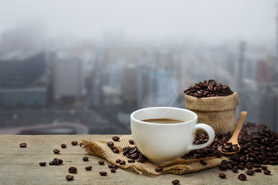A steaming cup of coffee sits on a wooden surface with scattered coffee beans, next to a burlap sack filled with more beans, against a foggy city skyline in the background.