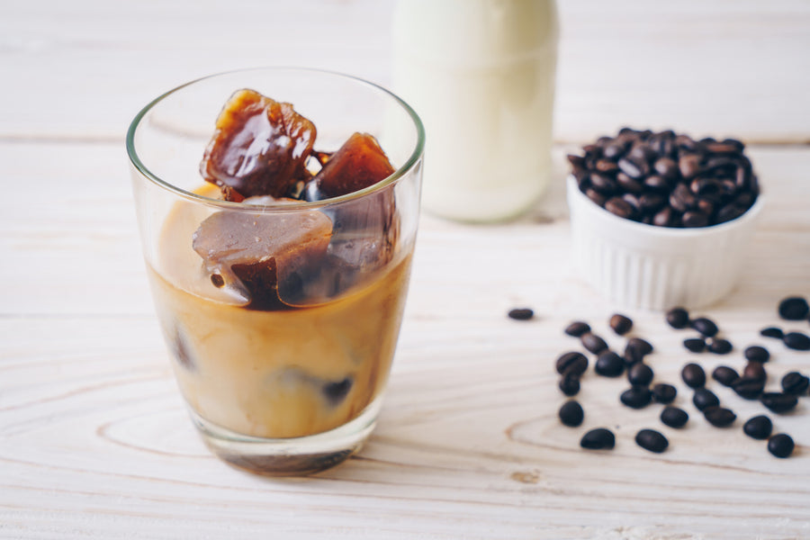 A glass of iced coffee with coffee ice cubes sits on a wooden table, accompanied by a bottle of milk and a bowl of whole coffee beans.