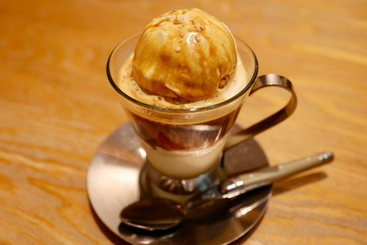 A clear glass cup filled with layered coffee and topped with a scoop of ice cream sits on a silver plate, accompanied by a small spoon, against a wooden table background.