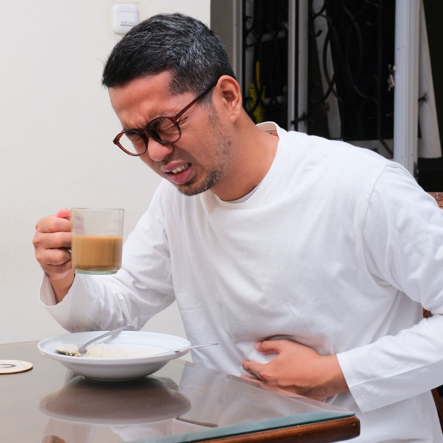 A man in a white shirt grimaces while holding a glass of coffee, sitting at a table with a bowl of rice, indicating discomfort in his stomach.
