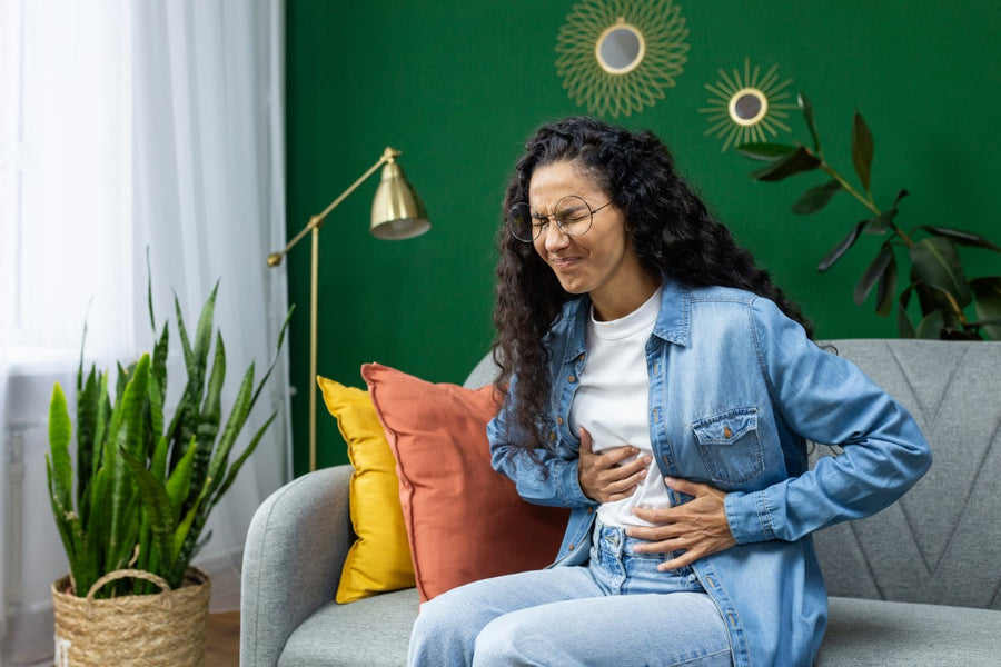 A woman with curly hair and glasses sits on a gray sofa in a green room, holding her stomach with a pained expression, surrounded by plants and colorful cushions.