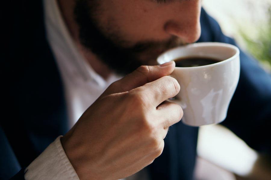 A man in a suit sips coffee from a white cup, with a focused expression, seated in a cozy cafe setting.