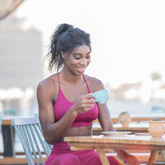 A woman in a pink athletic outfit smiles while holding a light blue cup, sitting at a wooden table outdoors with a blurred cityscape in the background.