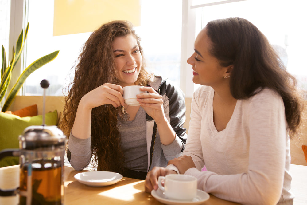 Two women sit at a table in a bright café, smiling and enjoying coffee together, with a French press and plates on the table and greenery in the background.
