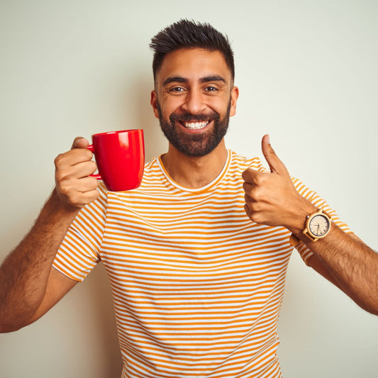 A smiling man with a beard holds a red mug in one hand and gives a thumbs-up with the other, wearing a striped shirt, against a light-colored wall.