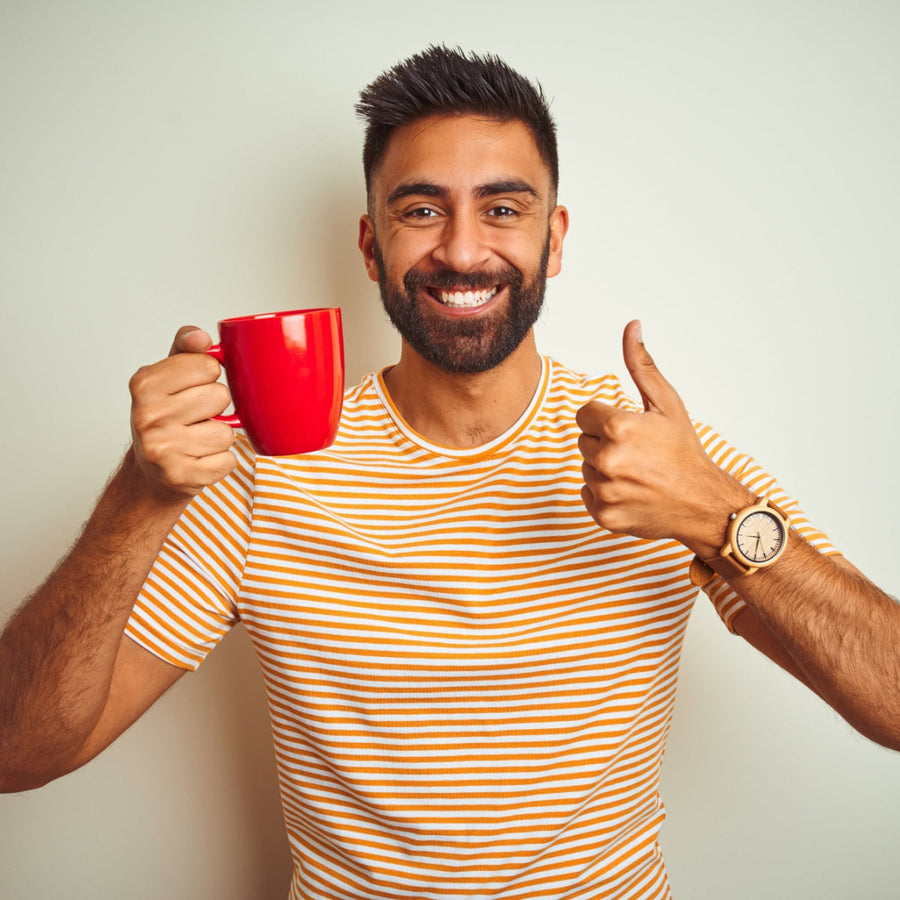 A smiling man with a beard holds a red mug in one hand and gives a thumbs-up with the other, wearing a striped shirt, against a light-colored wall.
