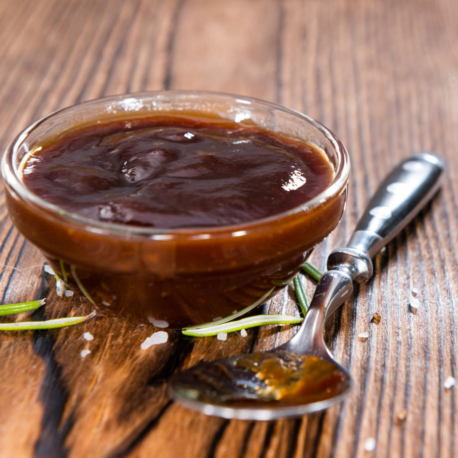 A glass bowl filled with thick, dark barbecue sauce sits on a wooden surface, accompanied by a silver spoon and scattered salt and rosemary leaves.