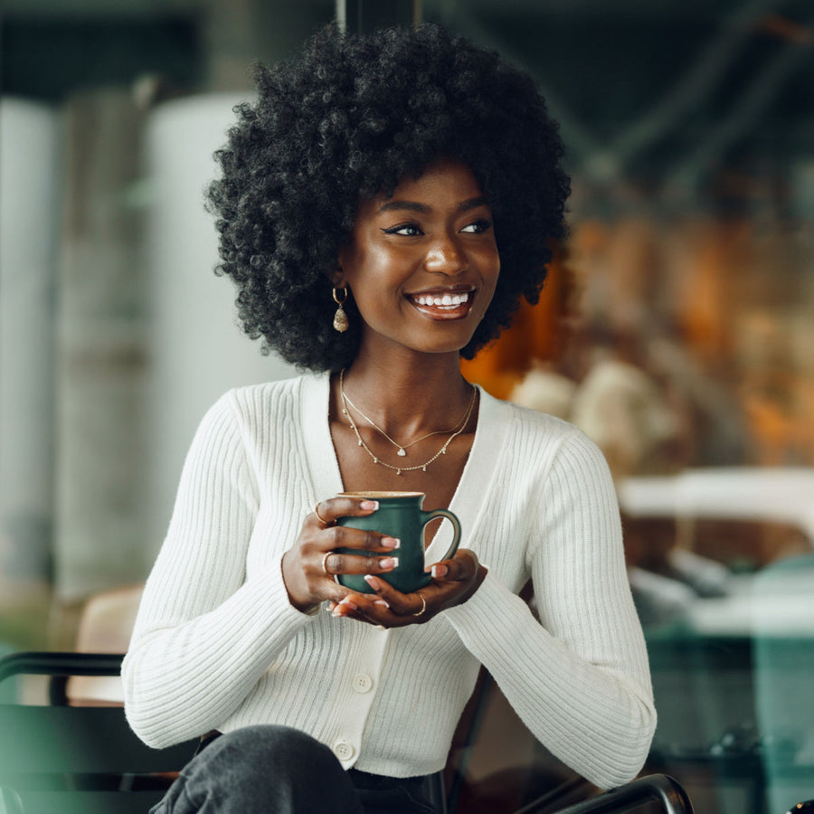 A woman with curly hair smiles while holding a green mug, seated in a cozy café with warm lighting and blurred background elements.