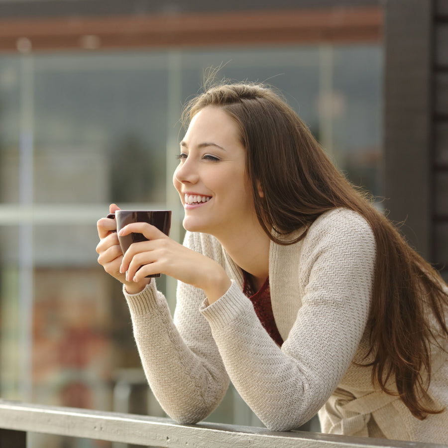 A smiling woman with long hair leans on a railing, holding a coffee cup, enjoying a moment outdoors in a bright setting with large windows in the background.