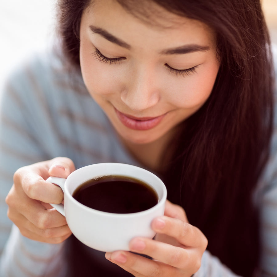 A young woman with long dark hair smiles gently as she holds a white cup of coffee close to her face, enjoying the aroma in a cozy indoor setting.