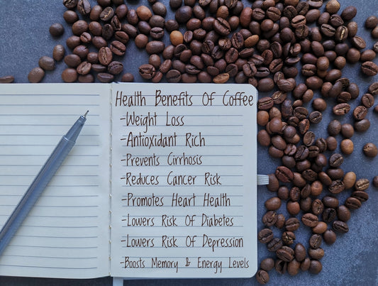 A notebook with a list of health benefits of coffee, including weight loss and heart health, sits on a dark surface surrounded by scattered coffee beans and a silver pen.