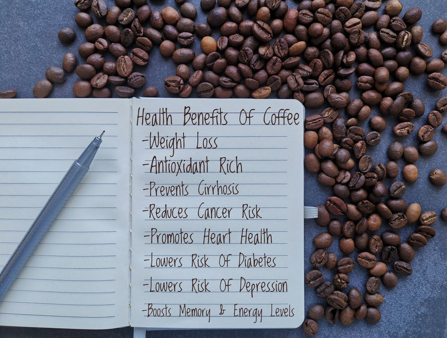 A notebook with a list of health benefits of coffee, including weight loss and heart health, sits on a dark surface surrounded by scattered coffee beans and a silver pen.