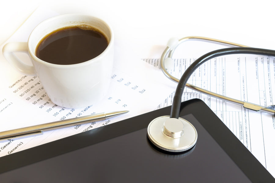 A white coffee cup sits next to a silver pen on medical paperwork, with a stethoscope resting on a tablet screen, suggesting a healthcare setting focused on patient care and documentation.
