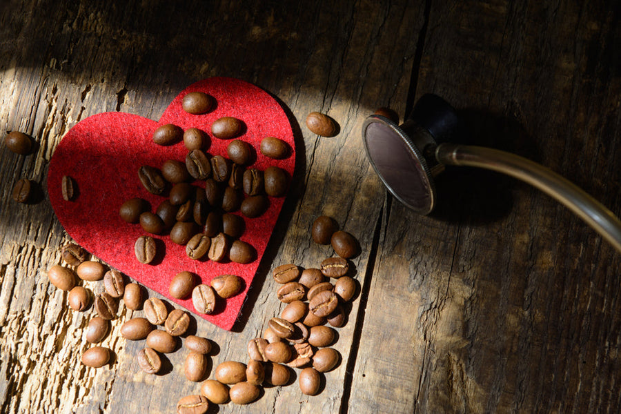 Brown coffee beans are scattered on a red heart-shaped felt piece, with a stethoscope lying nearby, all set against a rustic wooden background illuminated by soft light.