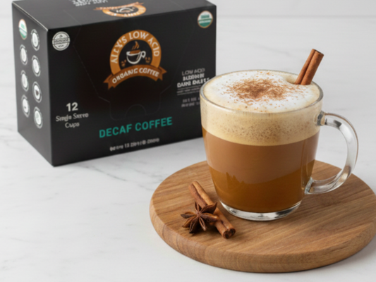 A clear glass mug filled with a layered cinnamon brown sugar latte topped with foam and cinnamon sits on a wooden board beside a box of low-acid decaf coffee.