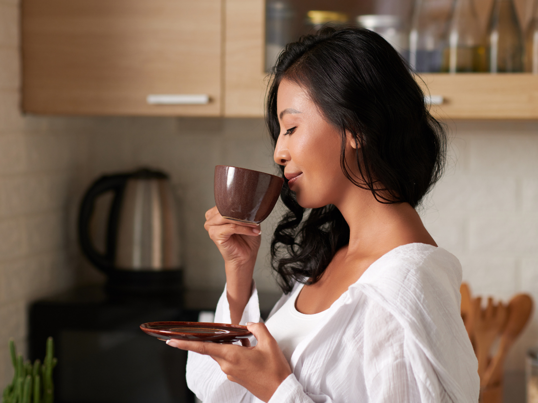 A woman in a white shirt enjoys a cup of coffee, savoring its aroma with a content expression, standing in a cozy kitchen with wooden cabinets and kitchenware in the background.