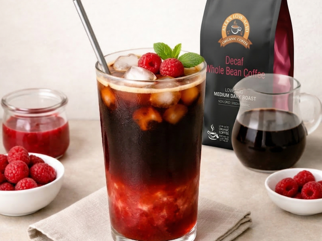 Iced coffee with raspberry syrup layered at the bottom, topped with fresh raspberries and mint, served alongside Alex’s Low Acid Decaf Whole Bean Coffee and additional coffee on a countertop.