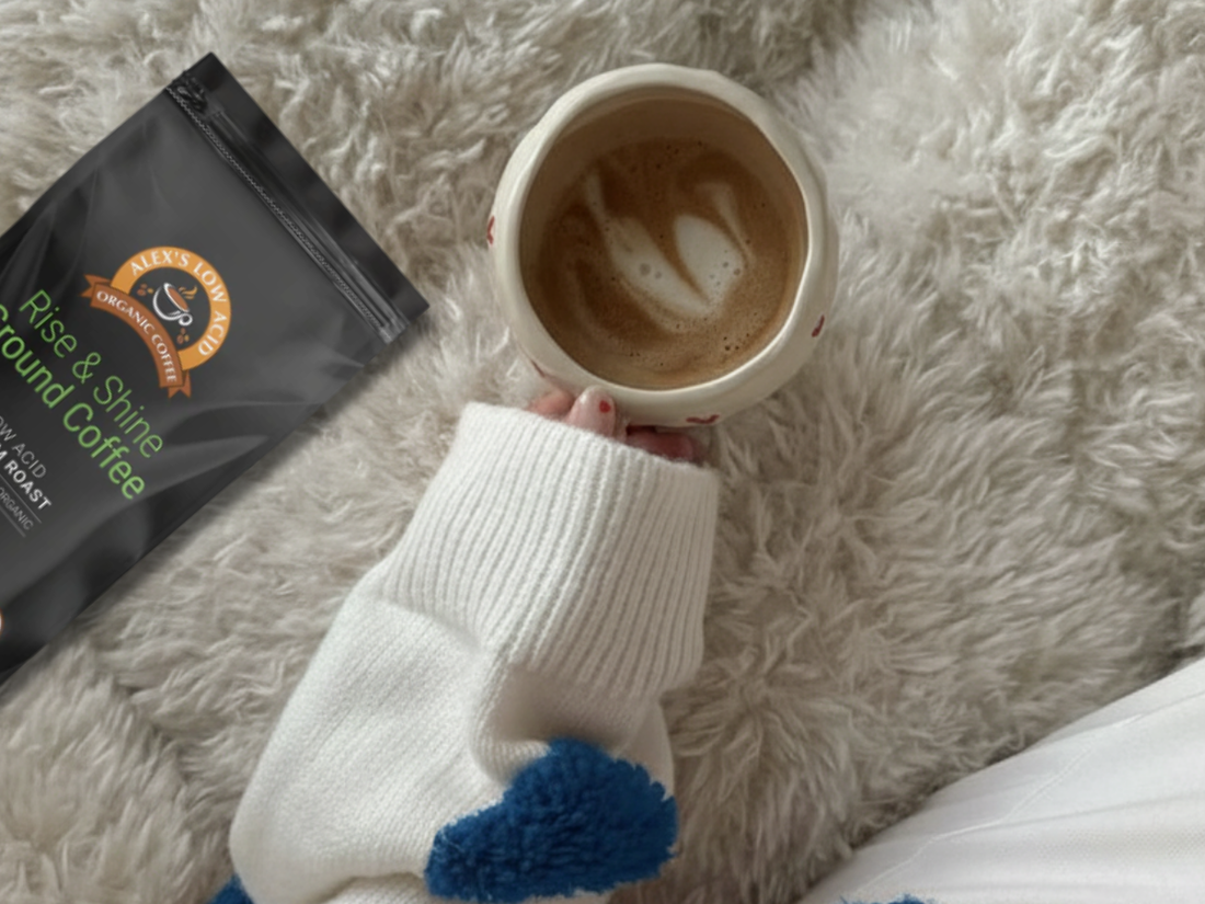 A hand in a cozy sweater holds a cup of latte with a heart-shaped foam design, resting on a fluffy blanket next to a bag of ground coffee labeled "Rise & Shine."
