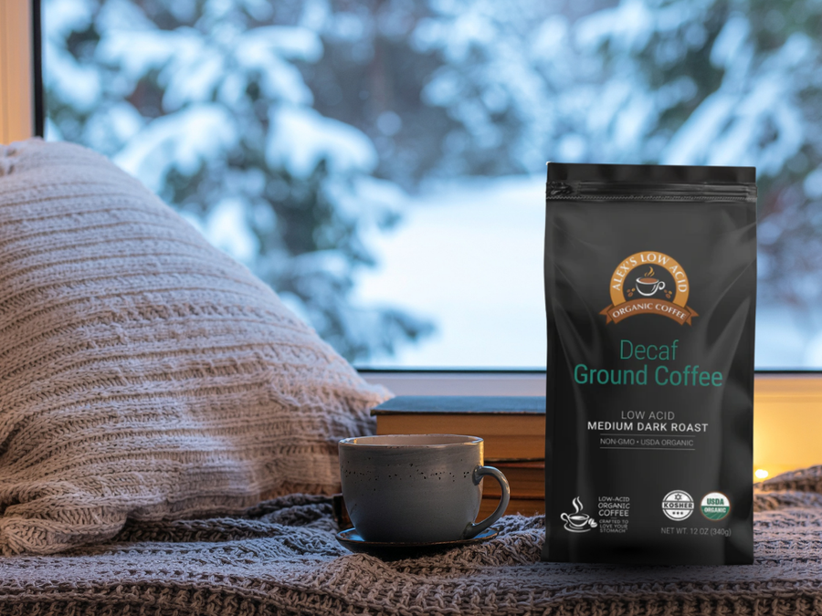 A cozy scene shows a cup of coffee on a wooden windowsill beside a bag of decaf ground coffee, with a snowy landscape visible outside and a soft, knitted pillow in the background.