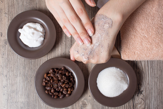 A hand applies a scrub to the other hand, surrounded by bowls of coffee beans, sugar, and cream, with a towel in the background, suggesting a DIY spa treatment.
