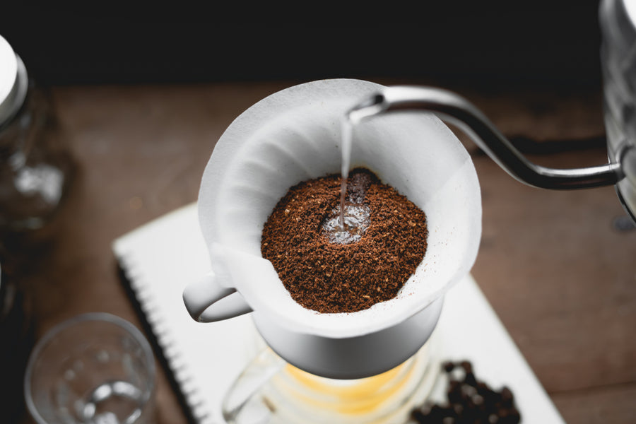 Hot water is being poured over ground coffee in a paper filter set in a white dripper, with a glass container below, surrounded by a wooden surface and a notebook.