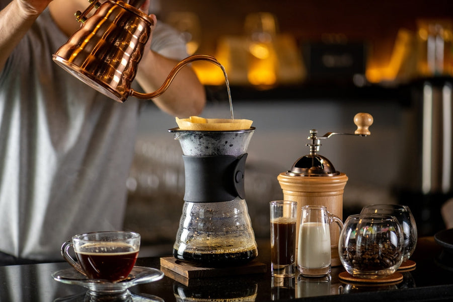 A person in a gray shirt pours hot water from a copper kettle into a coffee maker with a filter, surrounded by a cup of coffee, milk, a glass of espresso, and a coffee grinder on a dark countertop.