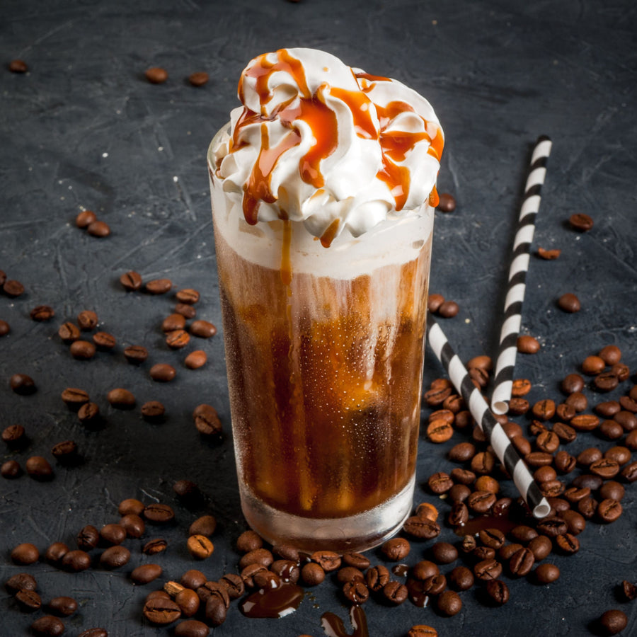 A tall glass filled with fizzy coffee topped with whipped cream and caramel drizzle sits on a dark surface scattered with coffee beans and two striped straws.