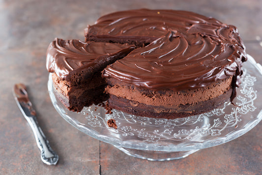 A rich chocolate cake with glossy frosting sits on a decorative glass cake stand, with a slice removed, revealing its layered texture against a rustic background.