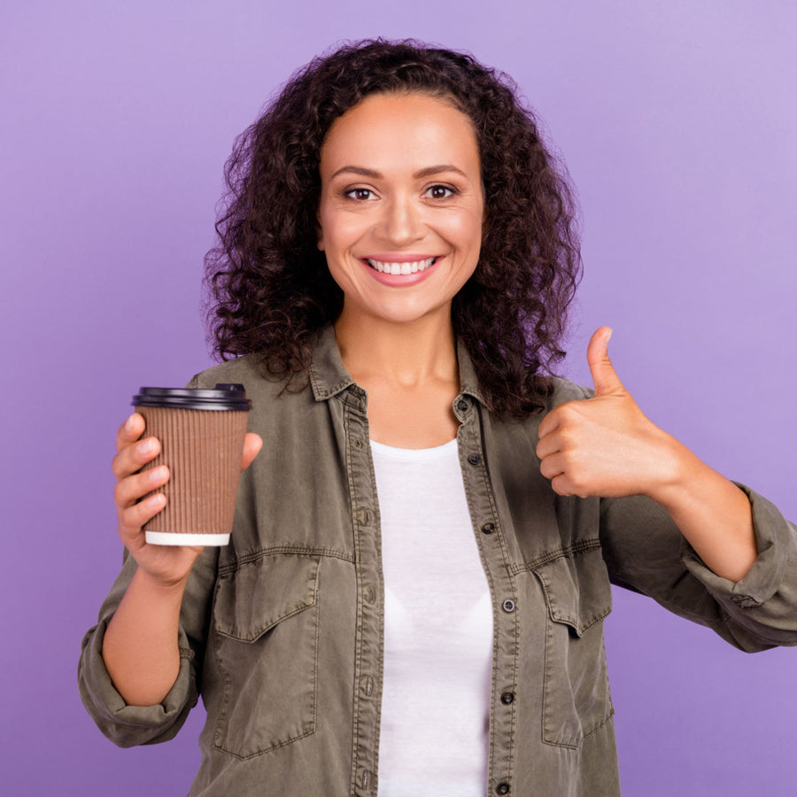 A smiling woman with curly hair holds a coffee cup in one hand and gives a thumbs-up with the other, set against a purple background.