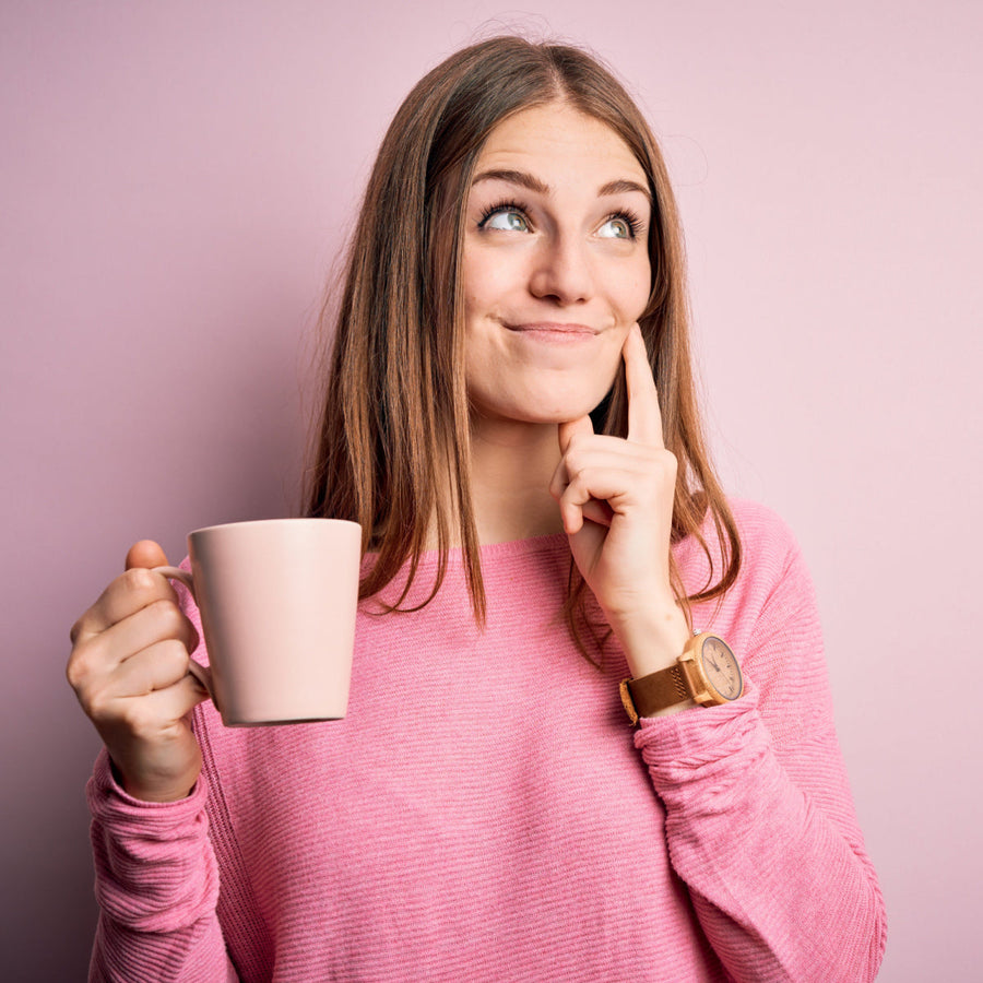 A young woman in a pink sweater holds a pink mug and smiles thoughtfully while resting her finger on her chin against a light pink background.
