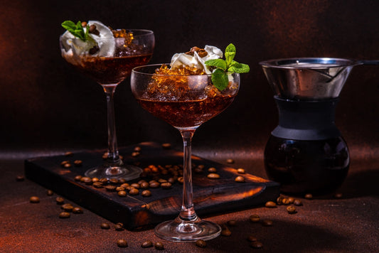 Two elegant cocktail glasses filled with dark liquid and topped with whipped cream and mint sit on a wooden board, surrounded by scattered coffee beans, with a coffee maker in the background.