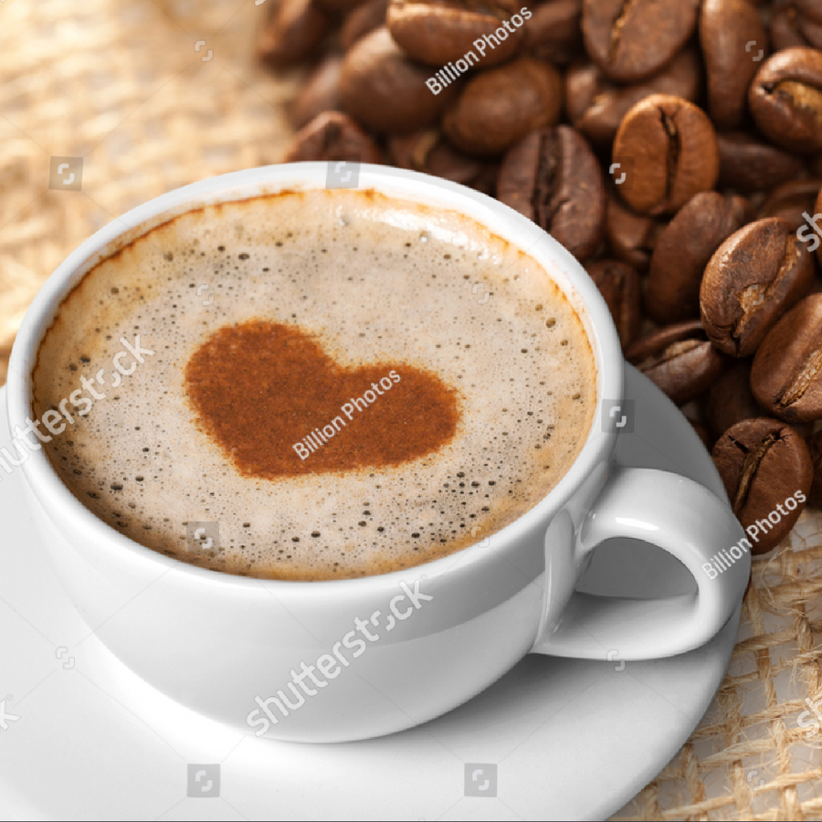 A white cup of coffee sits on a saucer, topped with frothy milk forming a heart shape, with roasted coffee beans scattered in the background on a textured surface.