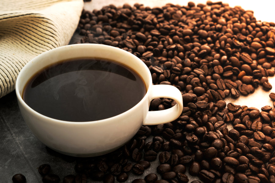 A white cup filled with steaming black coffee sits on a surface surrounded by scattered dark coffee beans, with a striped cloth partially visible in the background.