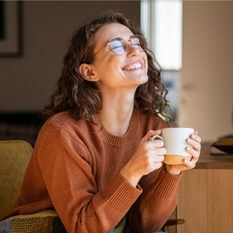 A woman with curly hair and glasses smiles joyfully while holding a white mug, sitting comfortably in a cozy living space with warm lighting.