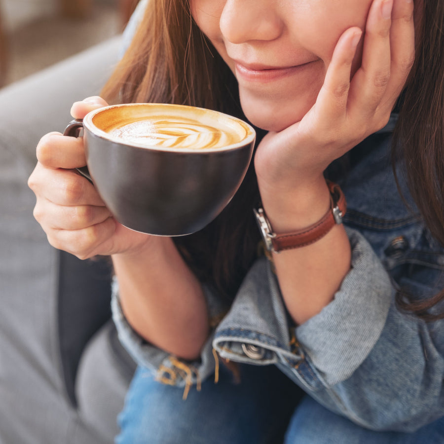 A woman with long hair smiles softly while holding a black coffee cup close to her face, sitting comfortably on a gray couch in a cozy setting.