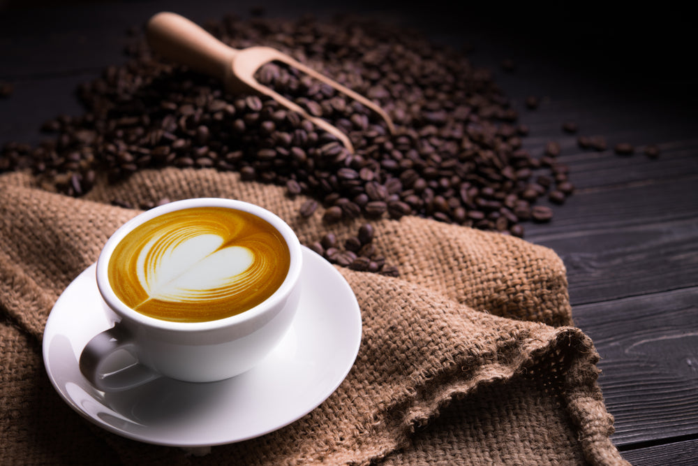 A white cup of latte with a heart-shaped foam design sits on a saucer, surrounded by scattered coffee beans and a burlap cloth on a dark wooden surface.