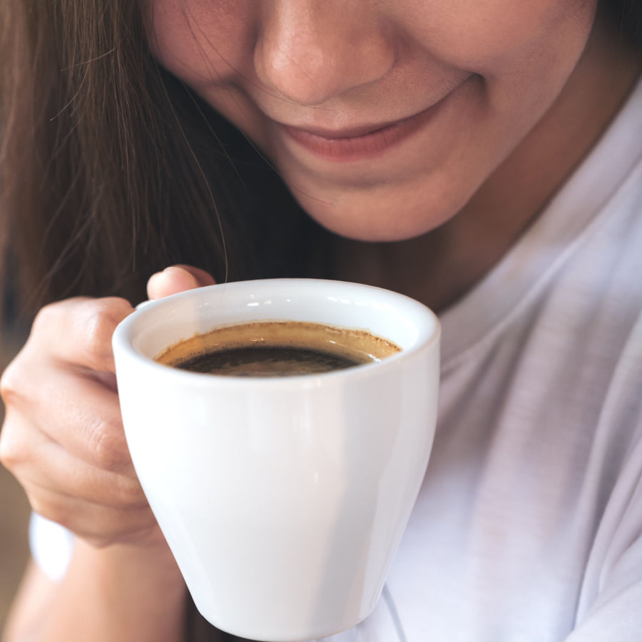 A woman smiles softly while holding a white cup of coffee close to her face, enjoying the aroma in a cozy indoor setting.